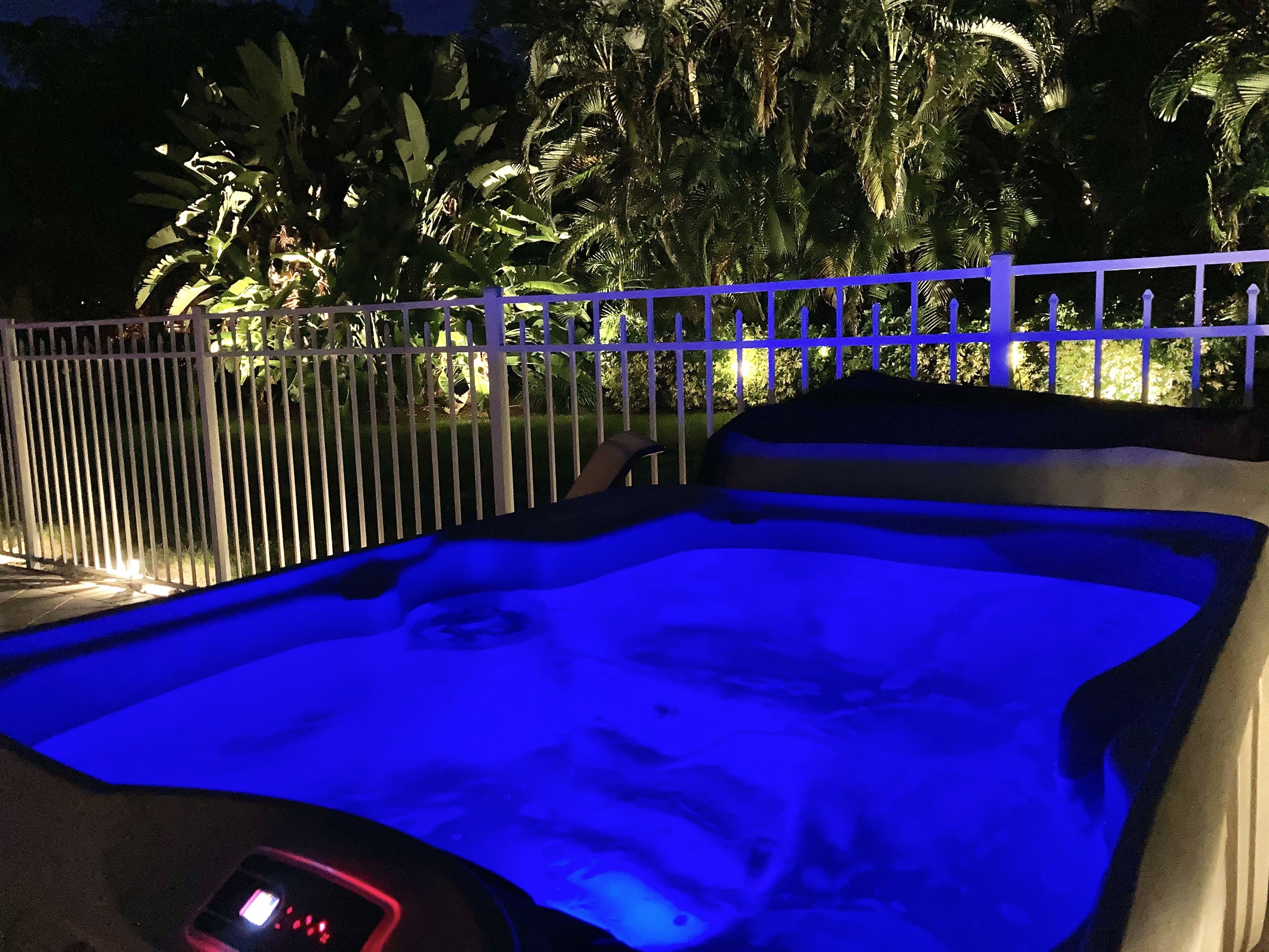 A hot tub with a blue light, surrounded by a white fence and palm trees.