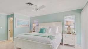 Une chambre avec un lit blanc, deux tables de chevet, un ventilateur de plafond et une fenêtre avec des stores.