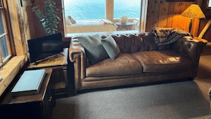 Et hyggeligt værelse med en lædersofa, et lille bord med en lampe og et vindue med udsigt over en strand.