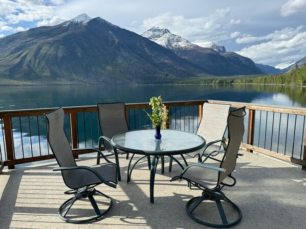 A deck with a round glass table and four chairs, overlooking a lake with mountains in the background. - Inside Glacier National Park, Lakefront cabin (Glacier National Park)
