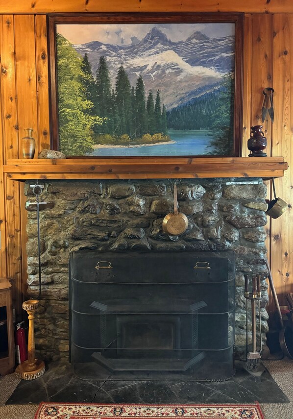 Una chimenea de piedra con hogar negro, una repisa de madera con un cuadro y decoración rústica que incluye un jarrón y una cuchara de madera.