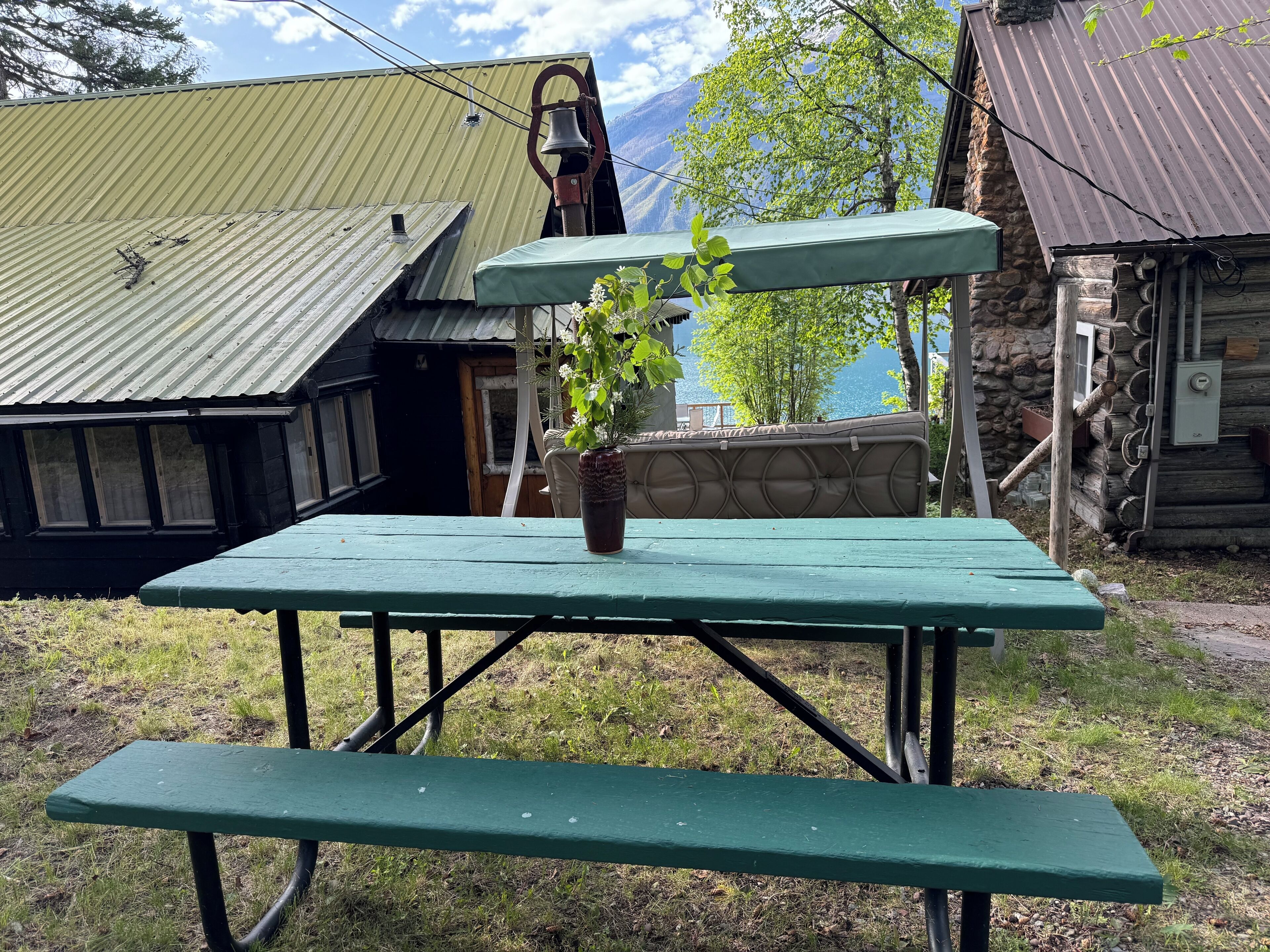 A picnic table with a potted plant in the center, surrounded by a stone wall and a wooden cabin with a metal roof.