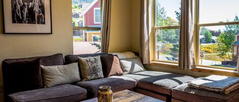 A cozy living room with a sectional sofa, a wooden coffee table, and large windows offering a view of a residential area.