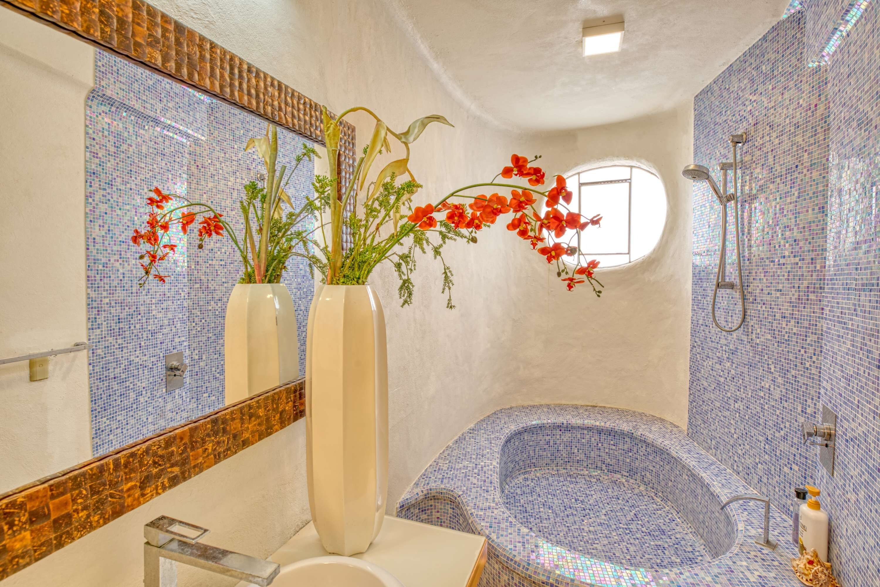 A bathroom with a mosaic-tiled bathtub, a large mirror, and two vases with flowers.