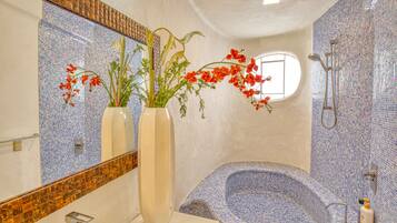 A bathroom with a mosaic-tiled bathtub, a large mirror, and two vases with flowers.