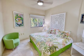 A bedroom with a bed, a green armchair, a window with a view of palm trees, and a decorative wall panel.