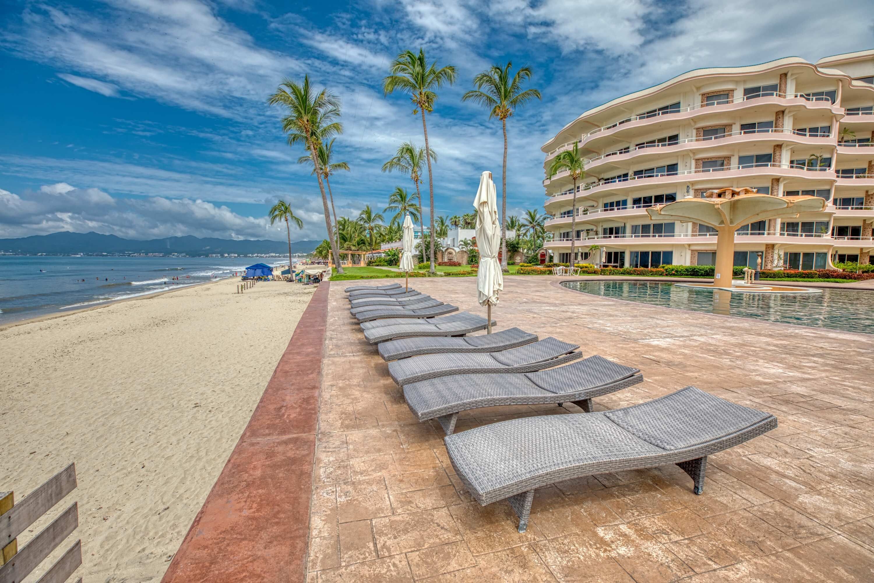 A beachside resort with a pool area, lounge chairs, and a large building with multiple balconies.