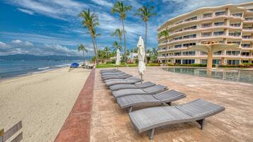 A beachside resort with a pool area, lounge chairs, and a large building with multiple balconies.