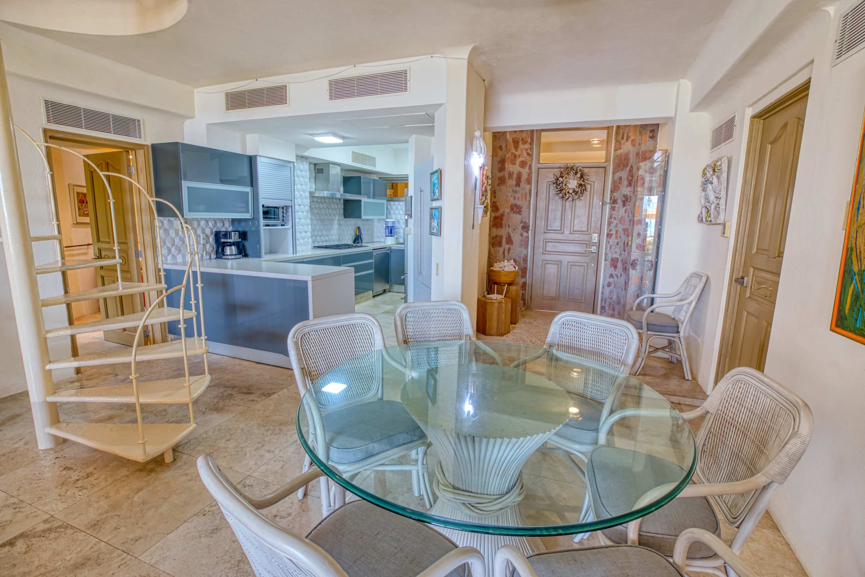 Une cuisine moderne avec des meubles blancs, un îlot central et un coin repas comprenant une table en verre et des chaises en osier.