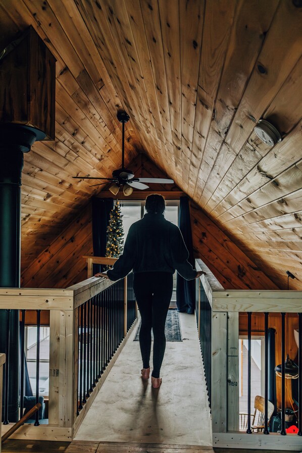 A person walking down a wooden staircase in a room with a sloped ceiling and a ceiling fan. - Ocean View • Hot Tub • Pet-Friendly Coastal Retreat (Parrsboro)