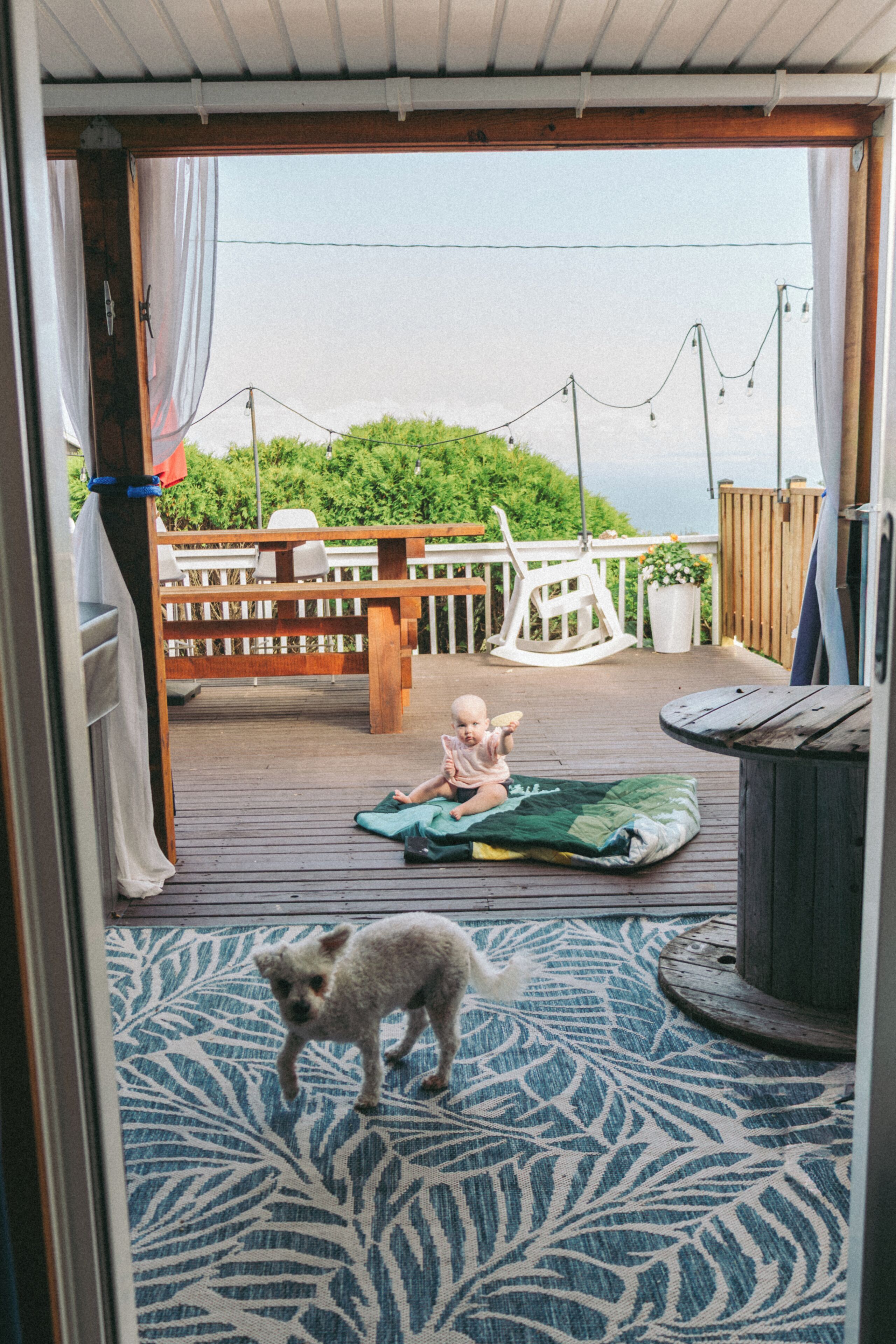 Una terraza de madera con un niño, un perro, una mecedora blanca y una valla de madera.