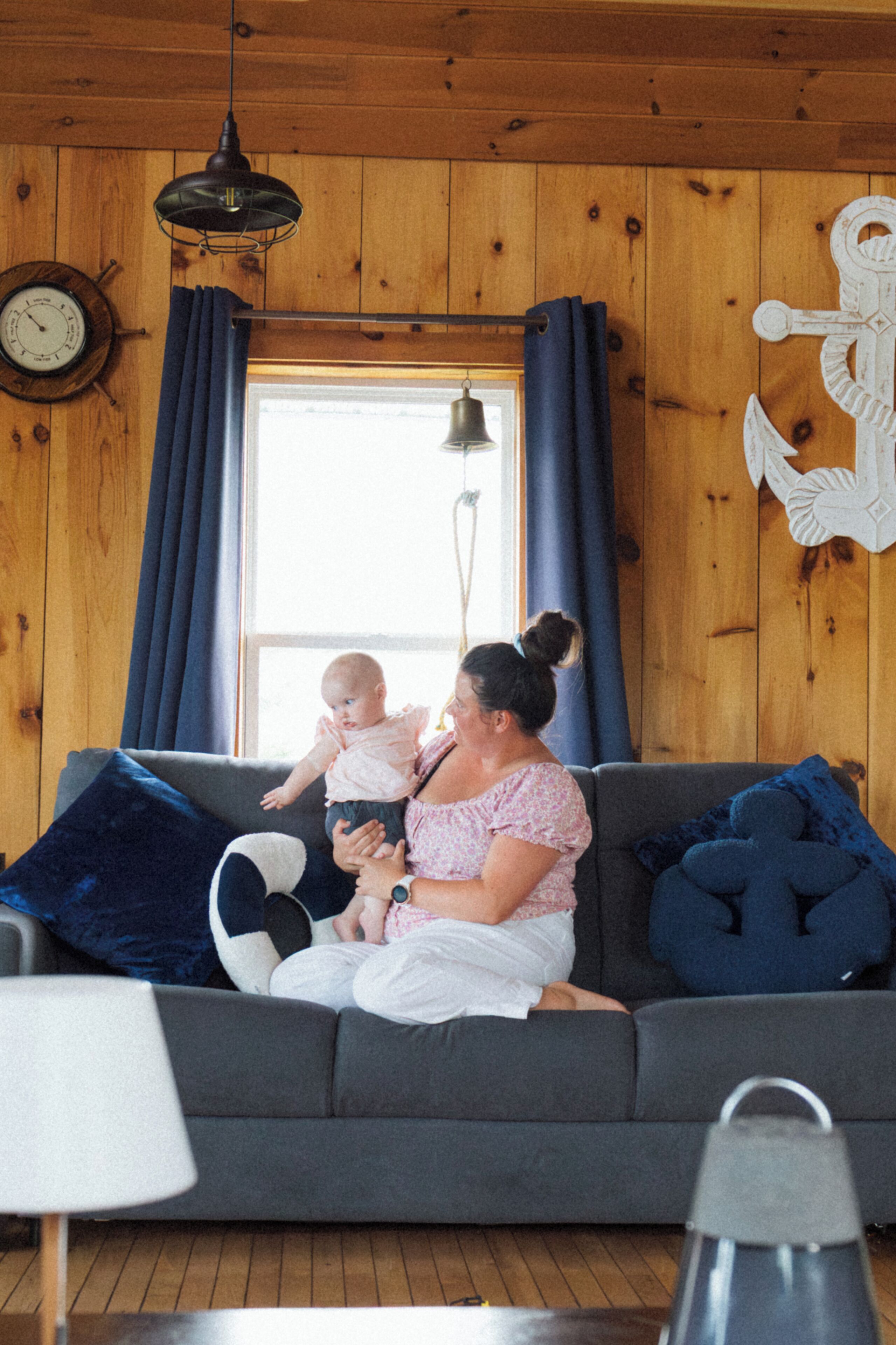 A room with a wooden interior, a sofa, a baby, and a woman sitting on the sofa.