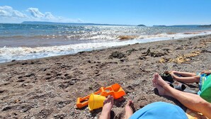 Een strand met een rotsachtige kustlijn, een kind dat in het zand speelt, en iemand in een groen shirt.
