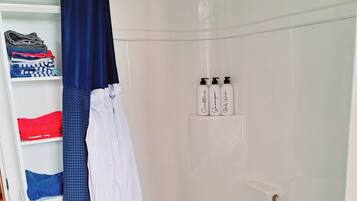 Un petit espace douche privé, avec un mur recouvert de carreaux blancs, un rideau de douche bleu et un banc. On y trouve trois bouteilles de shampoing et d’après-shampoing, posées sur le mur.