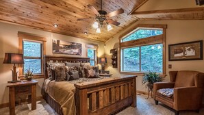 A bedroom with a wooden bed, a ceiling fan, large windows, and a view of trees.