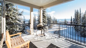A covered balcony with a wooden bench, a grill, and a view of snow-covered trees and a distant landscape.