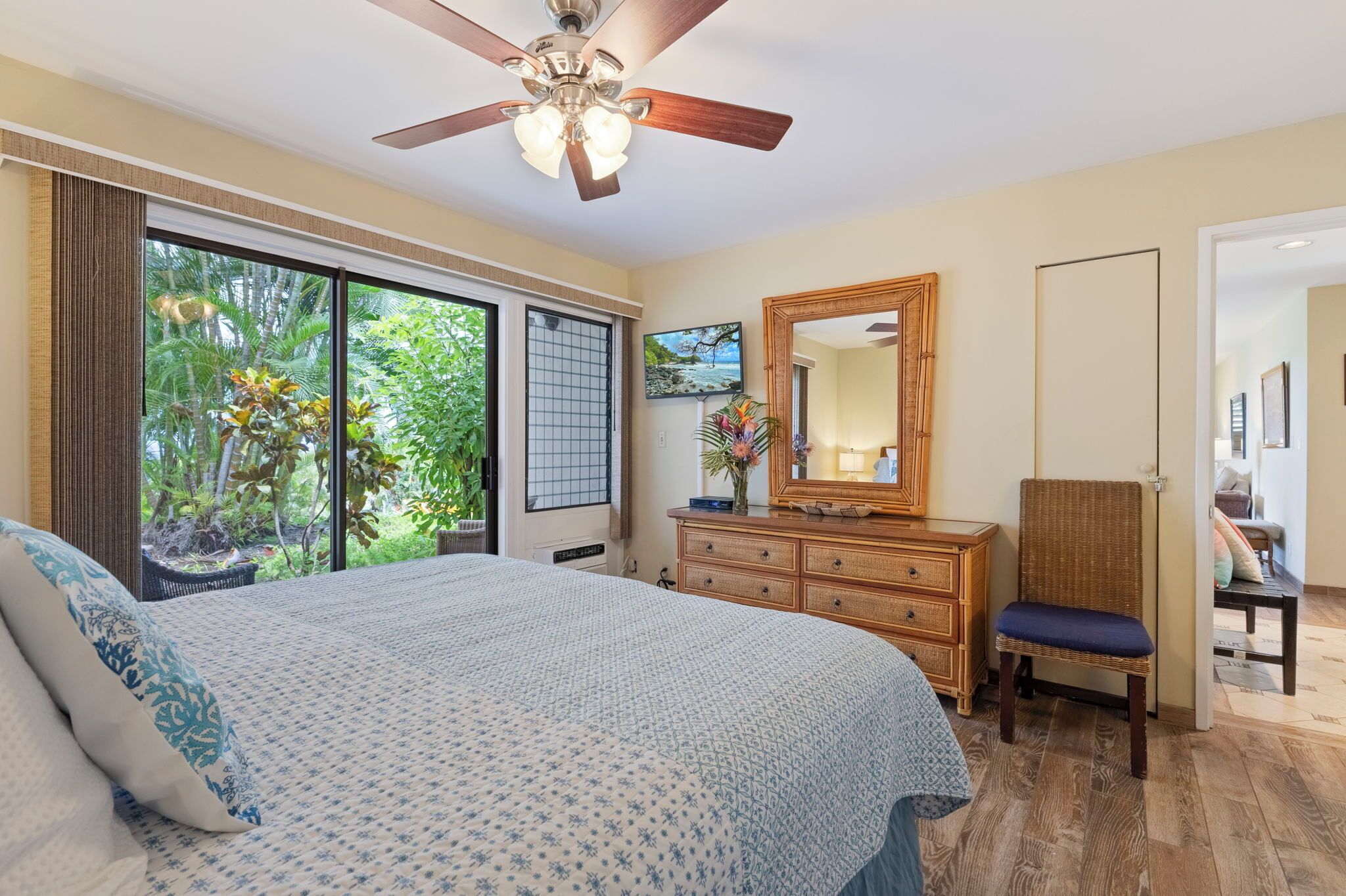 Un dormitorio con una cama grande, un aparador, una silla, un ventilador de techo y vistas al verde exterior.