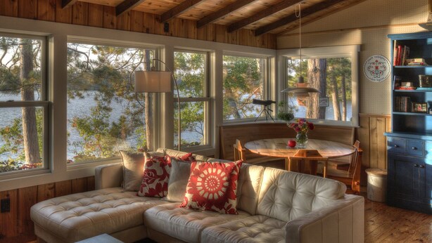 A cozy cabin interior with a wooden ceiling, large windows overlooking a lake, a cushioned sofa with patterned pillows, a dining table, and a bookshelf. - Classic Minnesota Cabin on Beautiful Lake O'Brien - Includes Pontoon Boat (Crosslake)