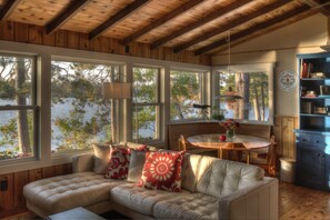 Ein gemütliches Cabin-Interieur mit Holzbalkendecke, großen Fenstern mit Seeblick, einer gepolsterten Couch mit gemusterten Kissen, einem Esstisch und einem Bücherregal.