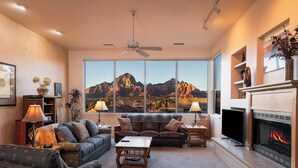 A living room with a large window, a fireplace, and a ceiling fan. - Spectacular Red Rock Views from Gorgeous Pueblo-style Sedona Home with hot tub! (Sedona)