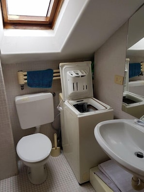 A compact bathroom with a skylight, a toilet, a washing machine, and a sink.