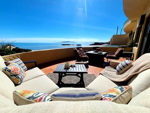 Un patio iluminado por el sol con vista al océano, amueblado con un sofá, sillas y una mesa.