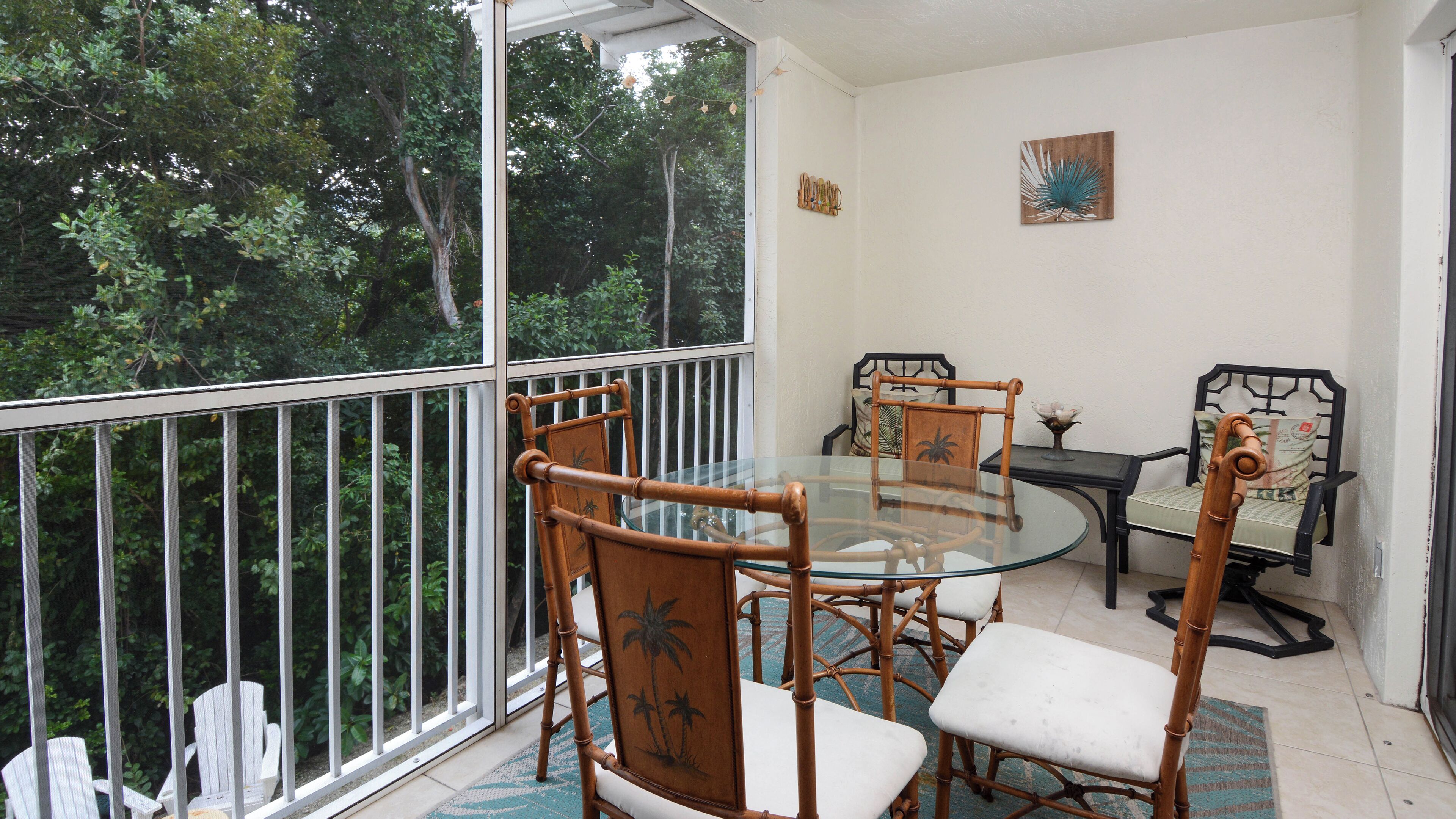 A balcony with a glass table and chairs, a view of trees, and a wall-mounted picture.