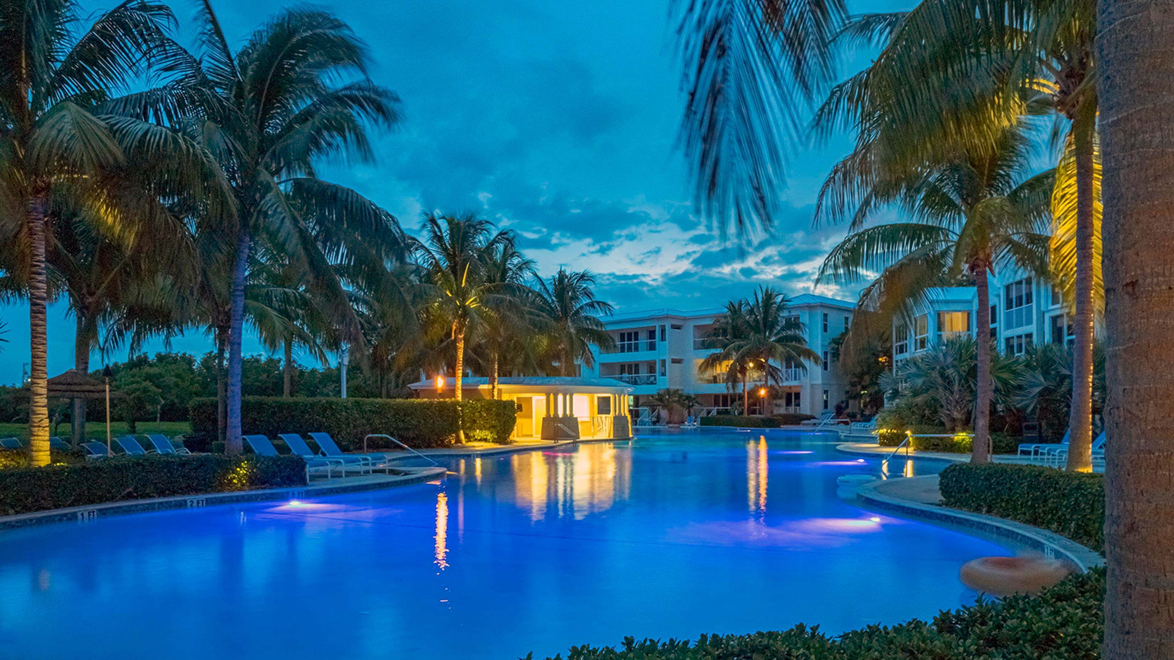 A swimming pool with palm trees, a building with balconies, and a clear sky.