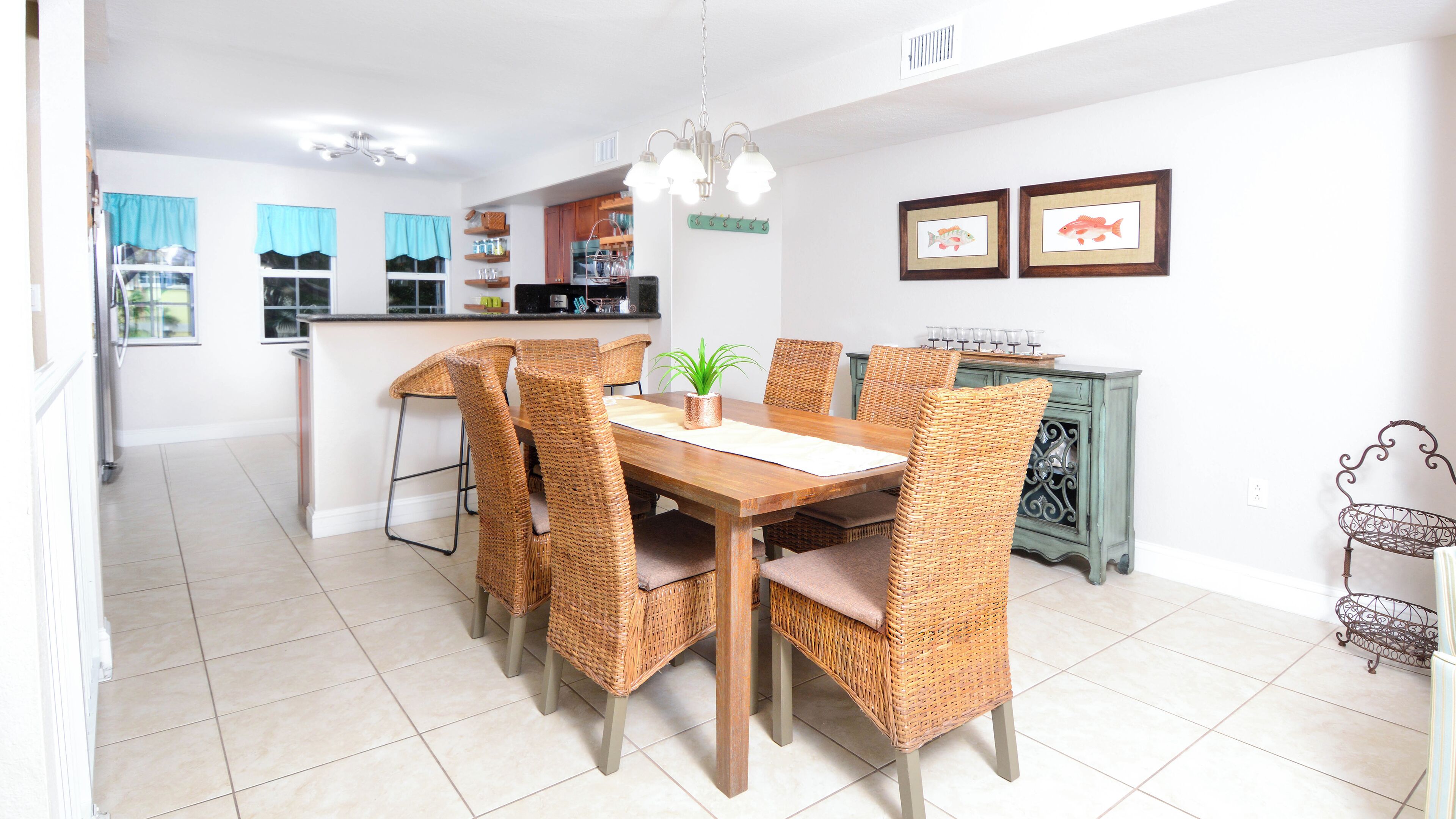 A dining area with a wooden table and wicker chairs, a kitchen in the background, and a decorative metal basket on the right.