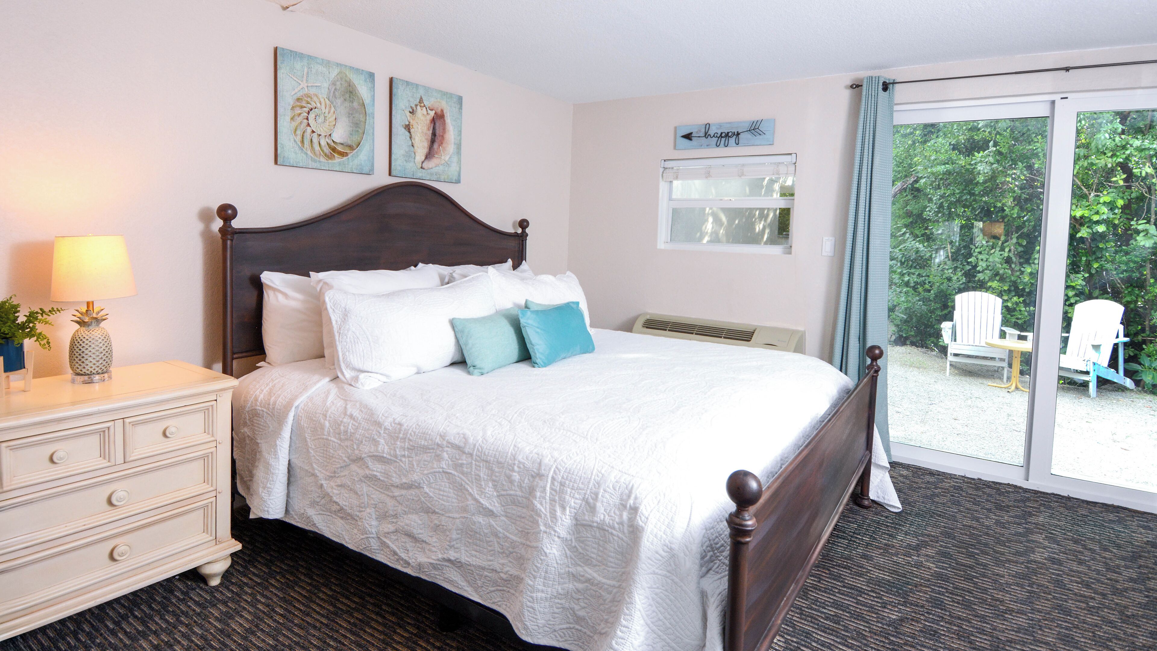 A neatly made bed with white linens and blue pillows, a wooden nightstand with a lamp, and a view of outdoor chairs through a glass door.