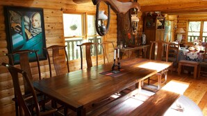 A wooden cabin interior with a large dining table, wooden chairs, a hanging light fixture, a painting on the wall, and a window with a view of trees. - PAPA BEAR'S LODGE -Best Log Cabin in Nevada City Sleeps up to 10.  (Nevada City)