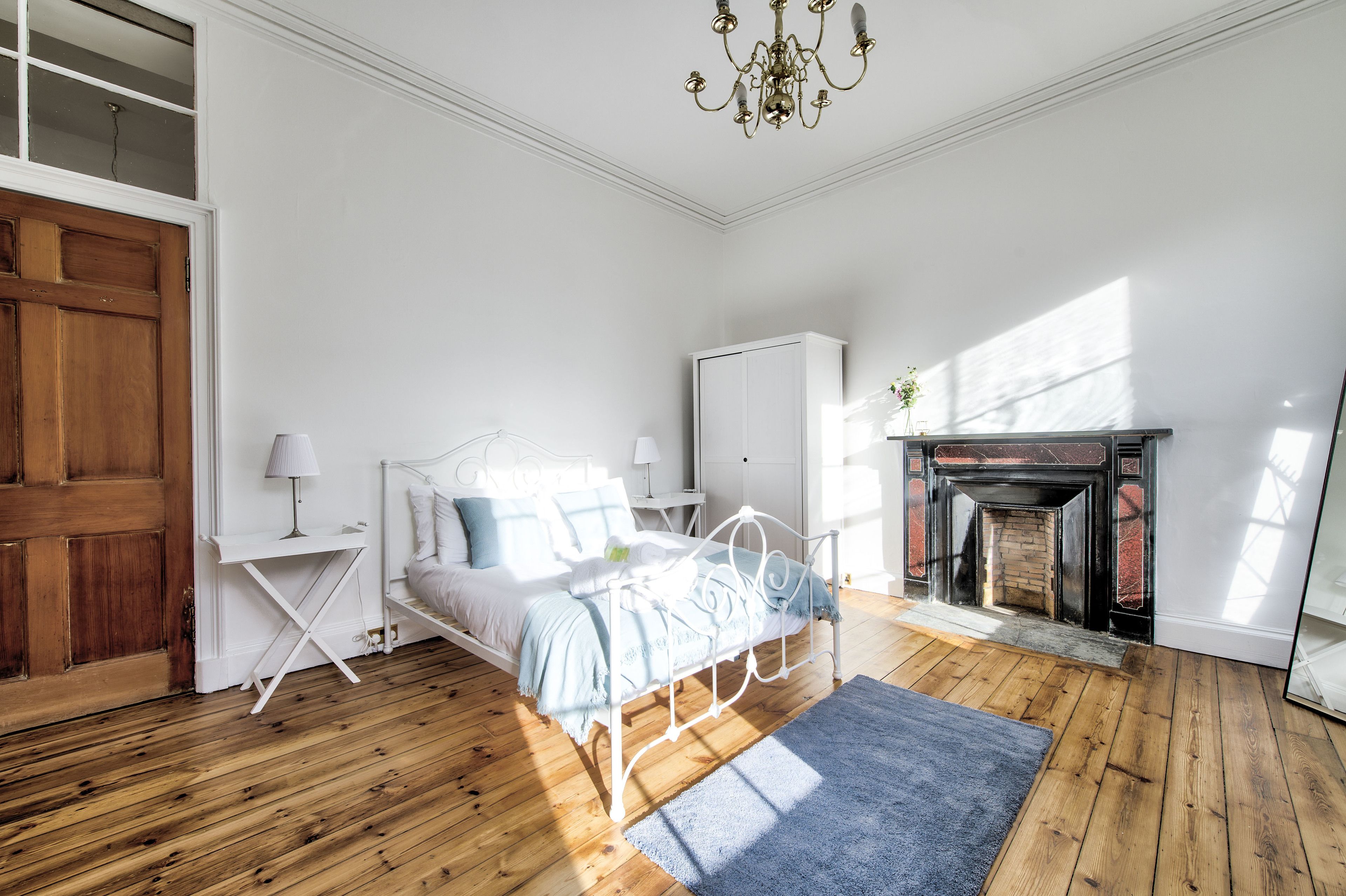 A bedroom with a wooden floor, a bed with white bedding, a bedside table with a lamp, a wardrobe, and a fireplace.