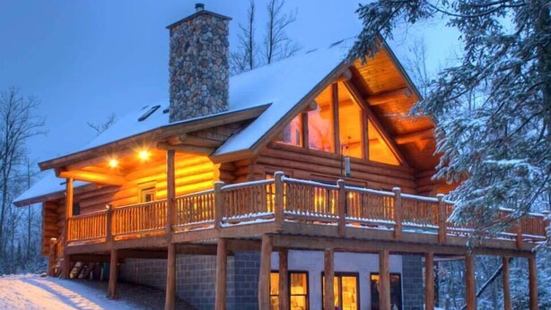 A log cabin with a stone chimney, wooden deck, and large windows.