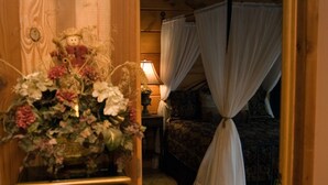 Un intérieur en bois agrémenté d’un arrangement floral posé sur un piédestal, un lit avec des rideaux blancs et une table de chevet avec une lampe.
