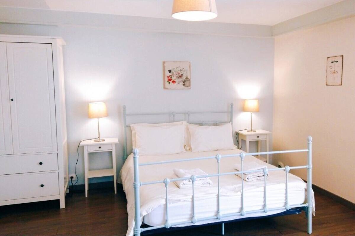 Un dormitorio con una cama de metal blanco, dos mesitas de noche con lámparas, un armario blanco y una obra de arte colgada en la pared.