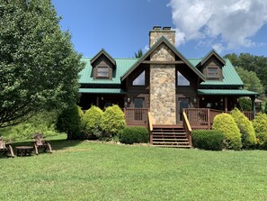 A large, two-story log cabin with a stone chimney, green roof, and a porch with wooden railings. - Wear's Valley Mtn Retreat - Pool across the Yard! Awesome Views, Easy Access (Sevierville)
