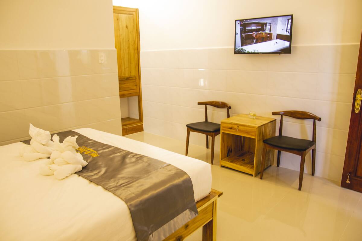 Una habitación de hotel con una cama, dos sillas de madera, una mesita pequeña y un televisor montado en la pared.