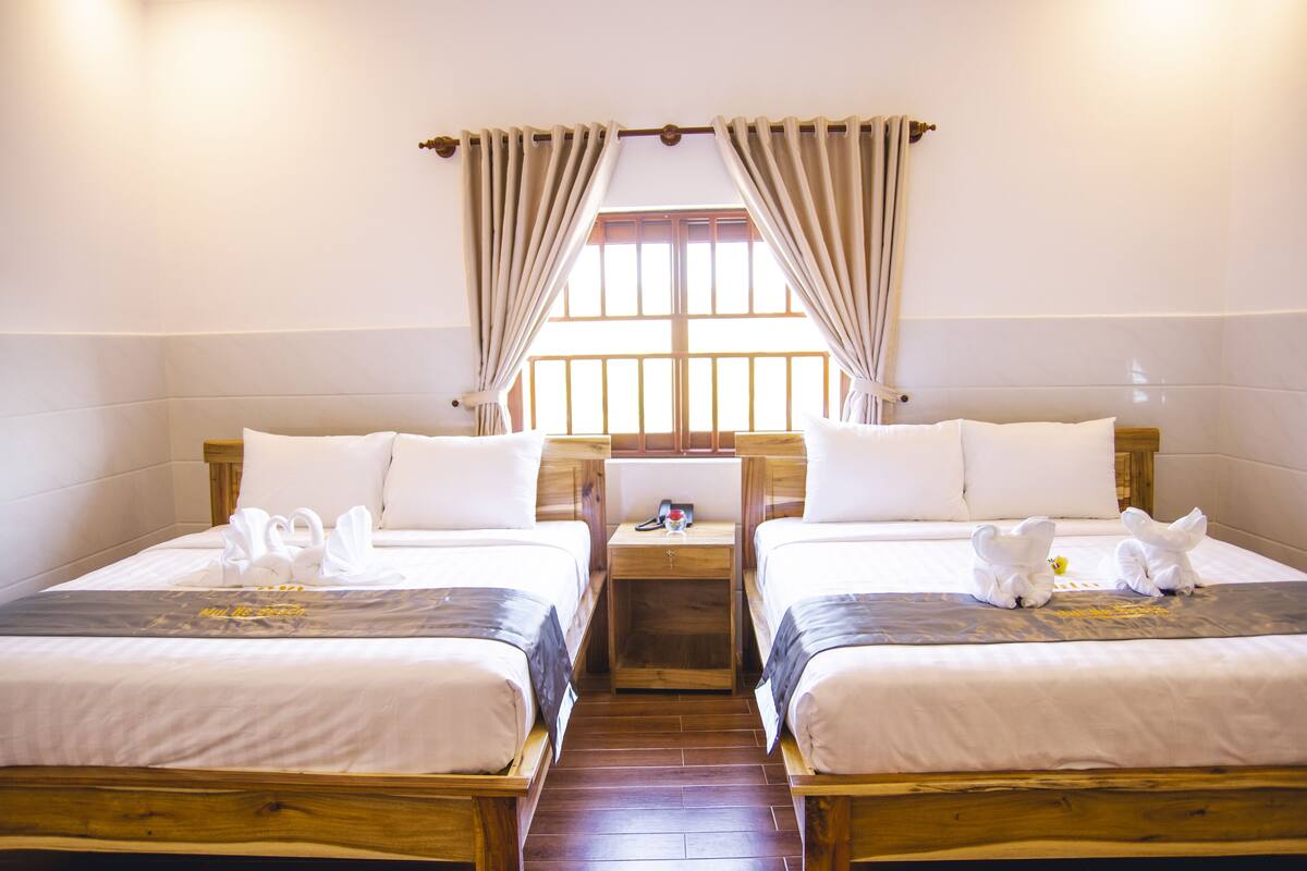 Habitación de hotel con dos camas individuales, cabeceros de madera, una mesita de noche con lámpara y ventana con cortinas.