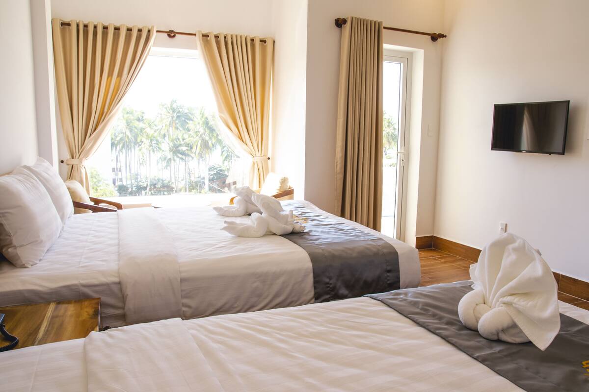 Habitación de hotel con dos camas, cortinas color beige, un televisor en la pared y vistas a palmeras.