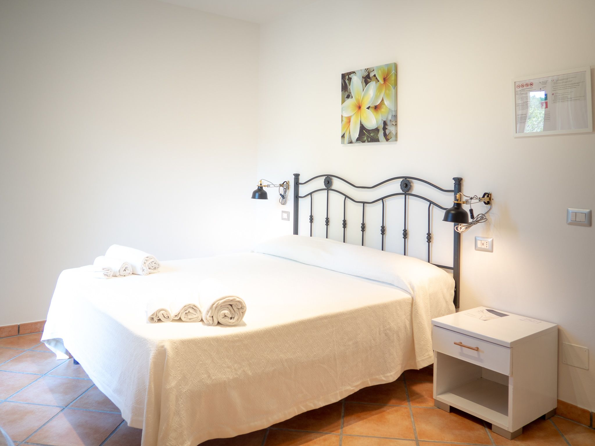 Een opgemaakt bed met witte lakens en opgerolde handdoeken, een zwart metalen hoofdbord, een nachtkastje met een lamp en een wanddecoratie van bloemen.