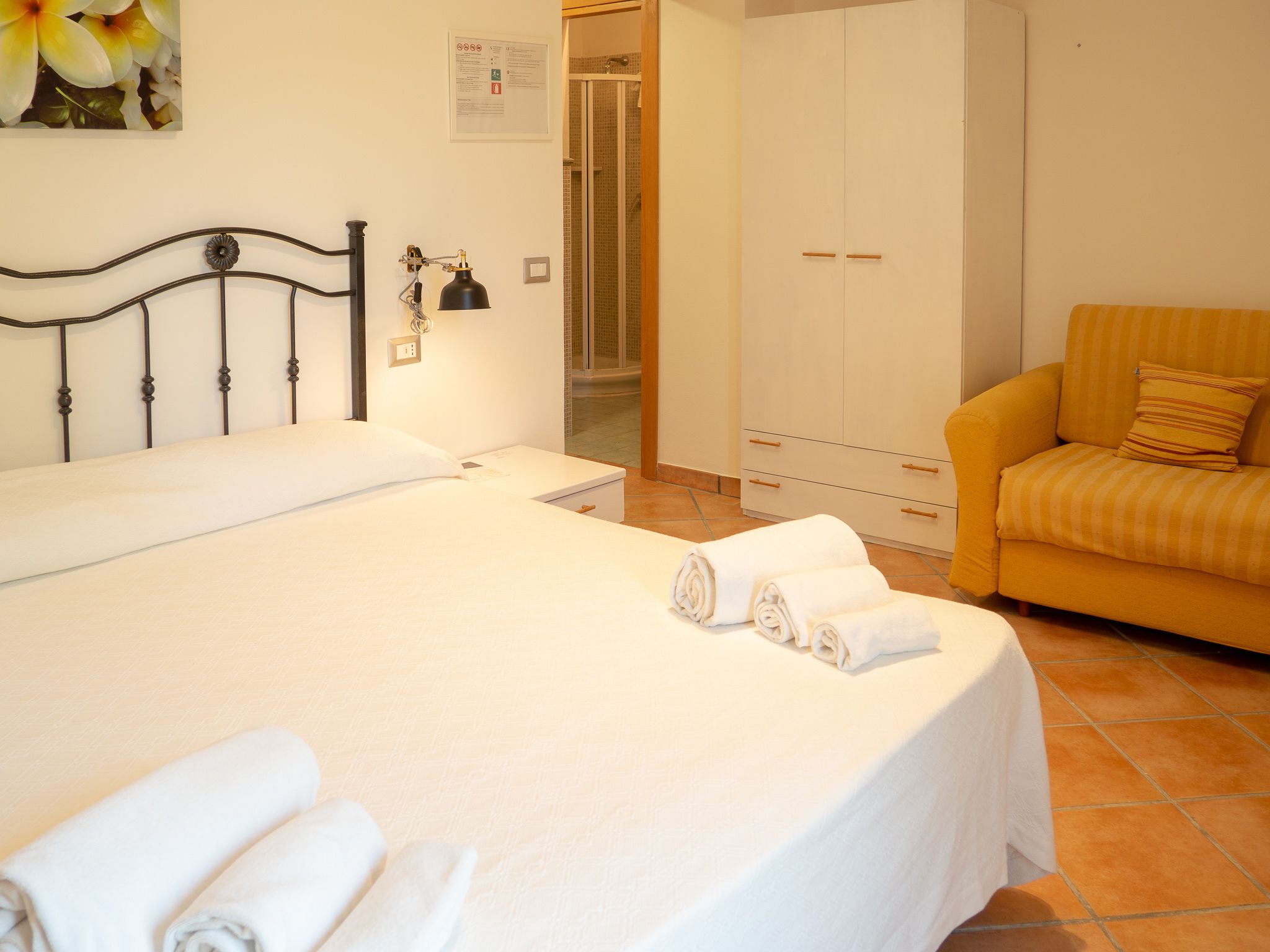 A neatly made bed with white linens and rolled towels, a black metal headboard, a white wardrobe with multiple doors, and a yellow-striped armchair.