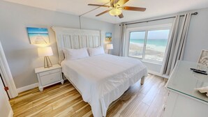 A bedroom with a bed, nightstands, a ceiling fan, a window with a beach view, and a glass table with a remote control.