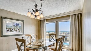 A dining area with a glass table and wooden chairs, a chandelier, a framed picture, and a view of the ocean through large windows.