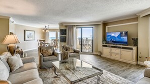 A spacious living room with a large flat-screen TV, comfortable seating, and a view of the ocean through the sliding glass doors.