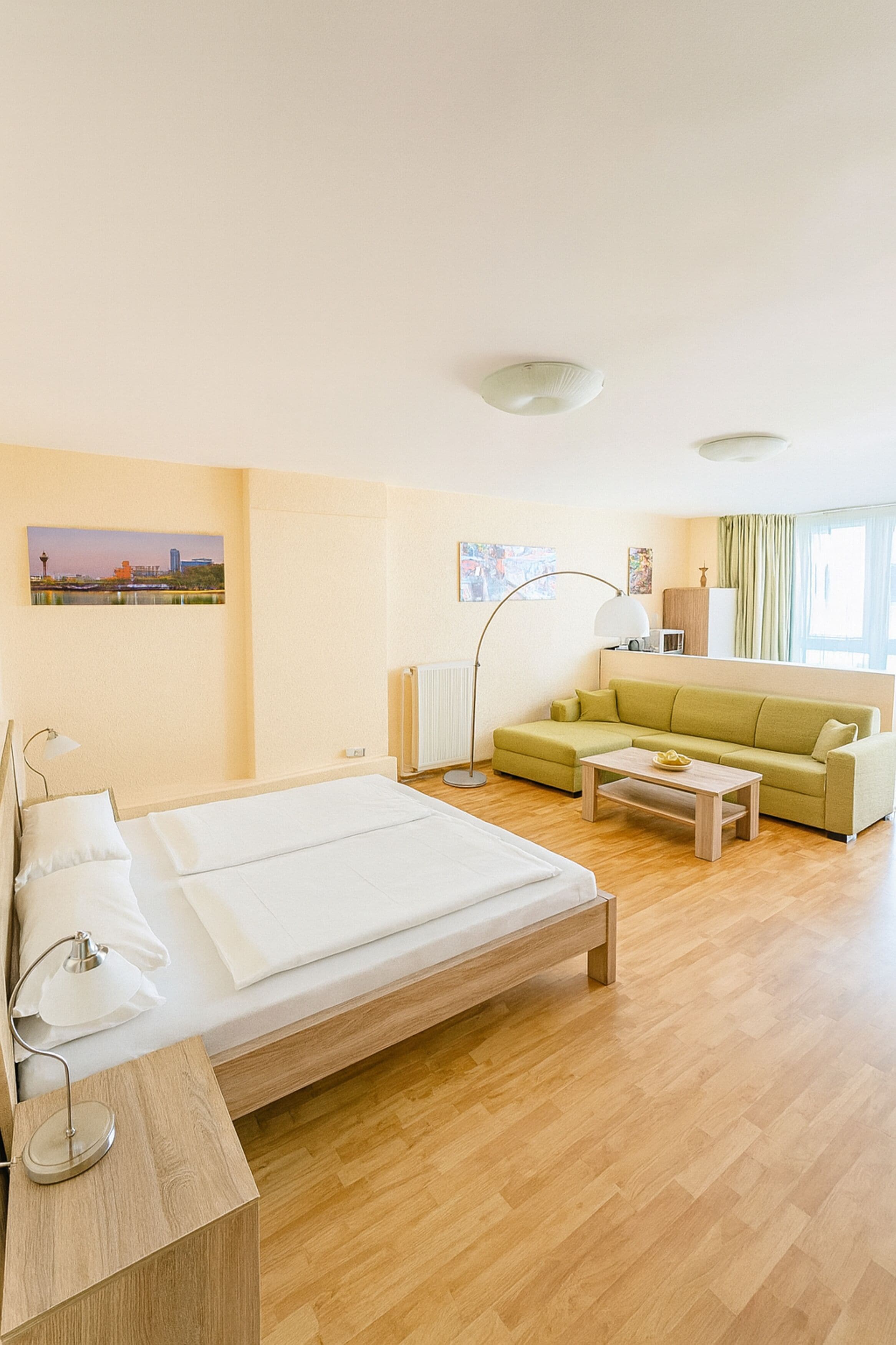 A hotel room with a bed, a green sofa, a wooden coffee table, a floor lamp, and a view of a cityscape.