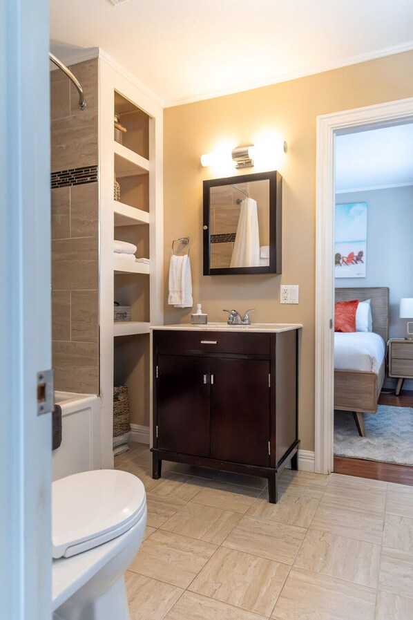 A bathroom with a toilet, a sink with a mirror, a shower, and a bed visible through an open door. - Cosy Oceanfront Cottage - Hot Tub and Sauna (South Shore)