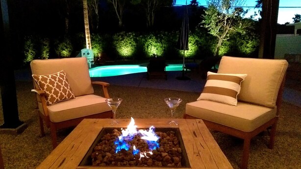 Two beige armchairs with patterned cushions, a square wooden table with a fire pit in the center, and a pool in the background. - A Mid-Century Modern Alexander Home Oasis (Lively Music-Themed Decor). (Palm Springs)