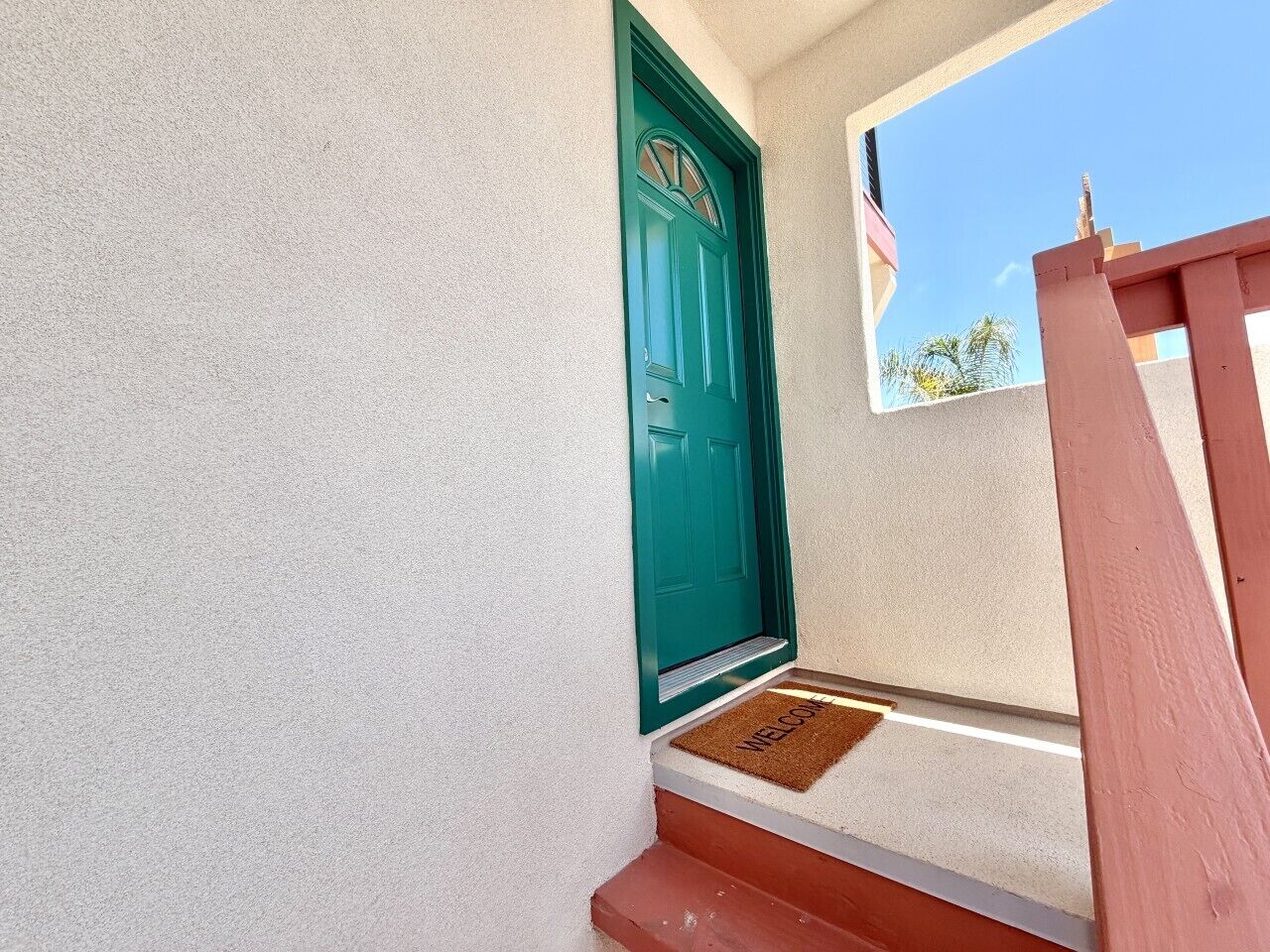 Una puerta azul verdoso con felpudo, una pared blanca con textura y un barandal de madera.