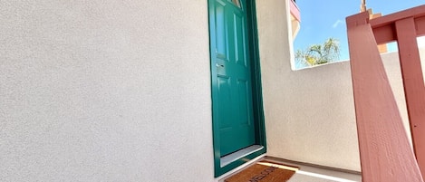 Una puerta azul verdoso con felpudo, una pared blanca con textura y un barandal de madera.