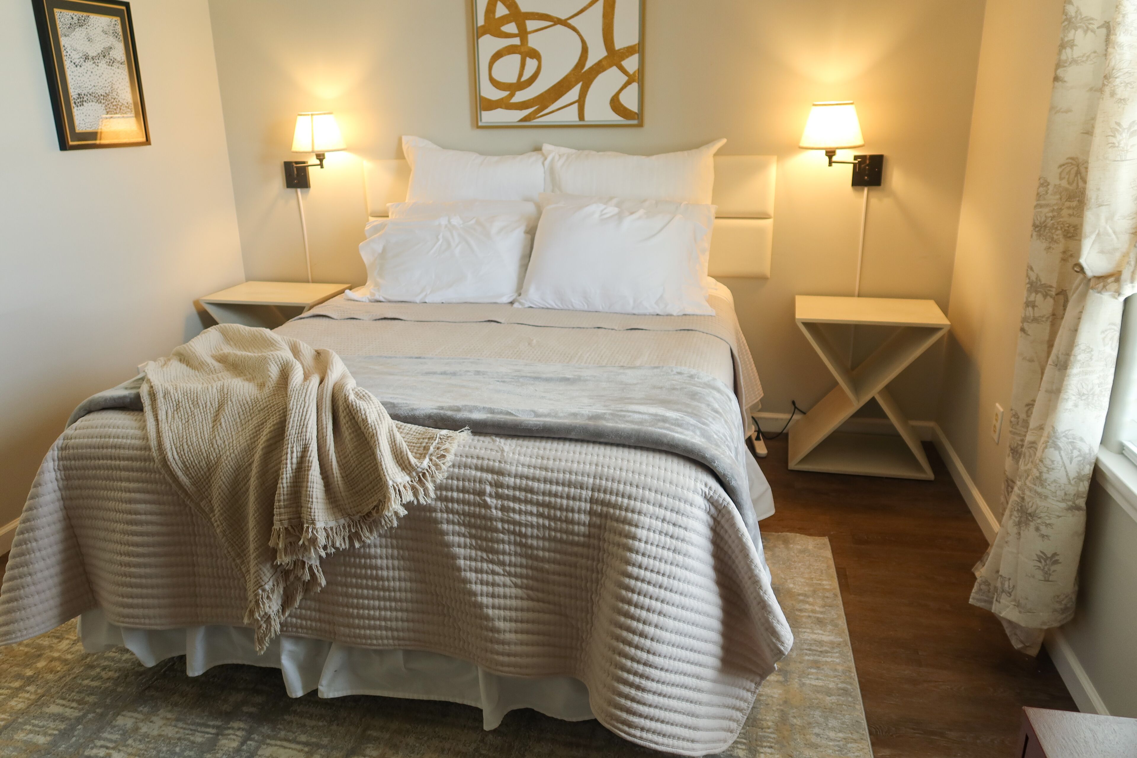 A neatly made bed with a beige blanket, two bedside lamps, and a wall art piece above the bed.